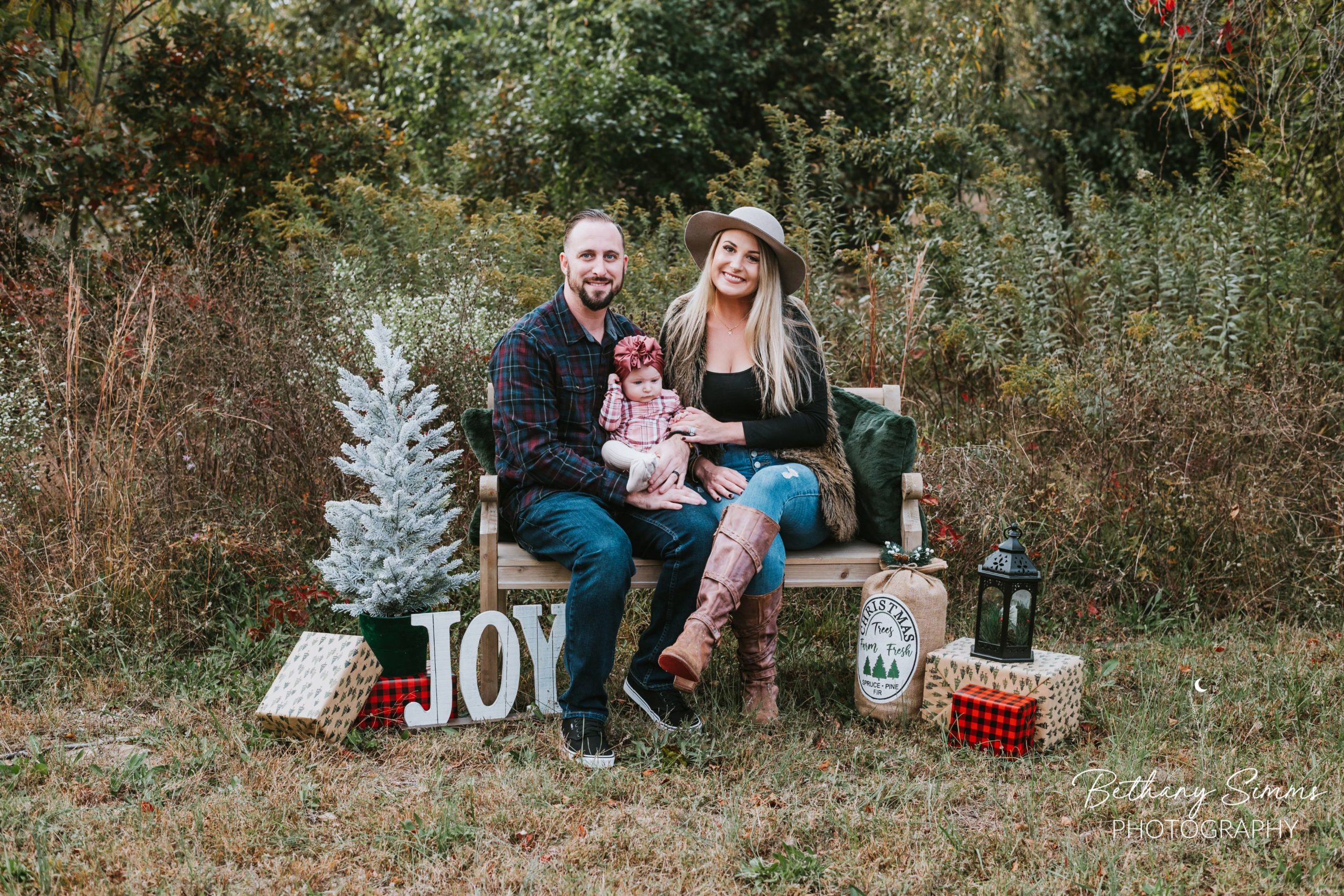 Mom, dad, and baby sitting on a bench with a Christmas tree, presents and other Christmas decorations for Christmas pictures mini session
