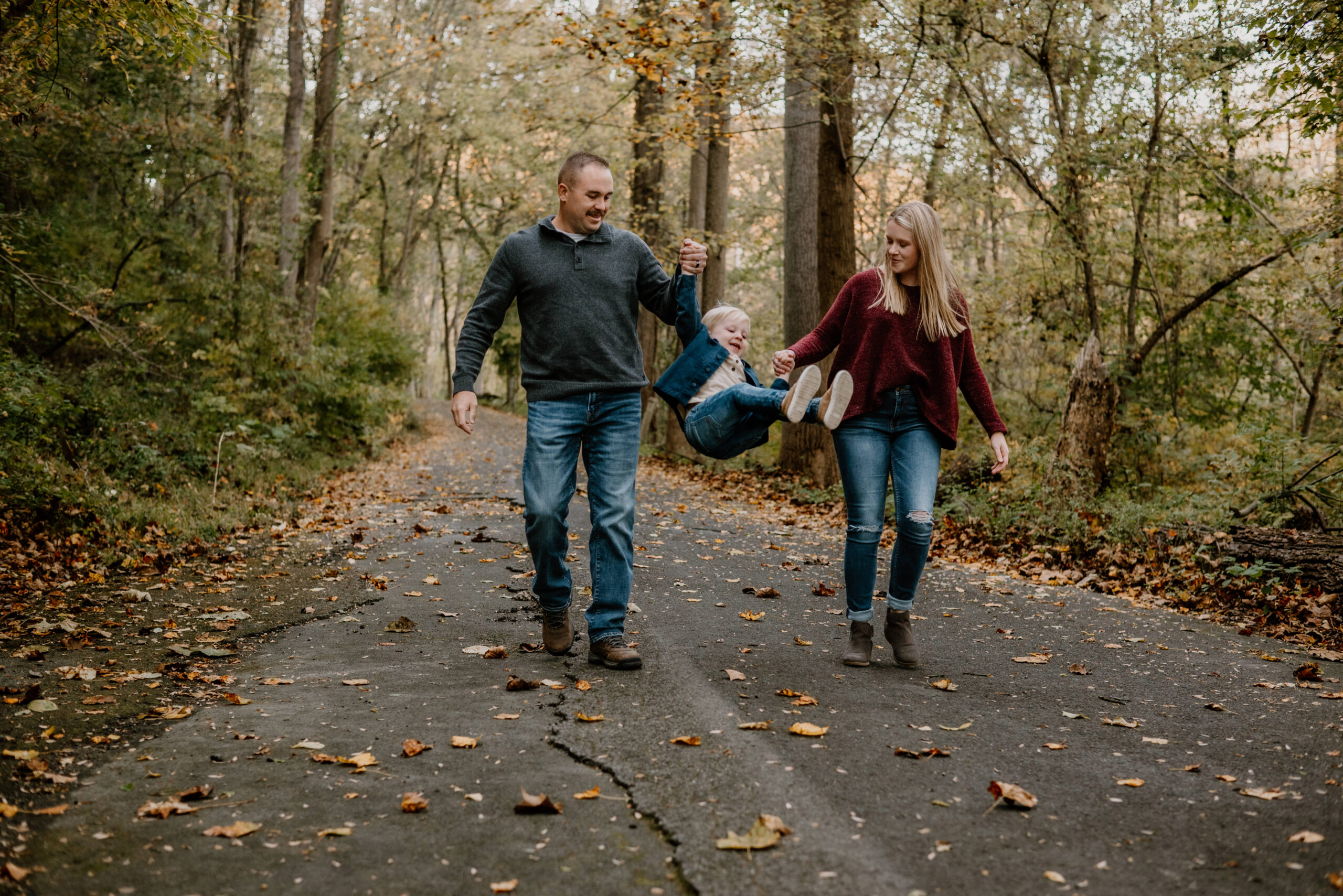 A family of three, dressed in cozy autumn attire, playing joyfully amid vibrant fall foliage in Baltimore, Maryland. They are smiling warmly, capturing the essence of a fun and memorable fall mini photography session.