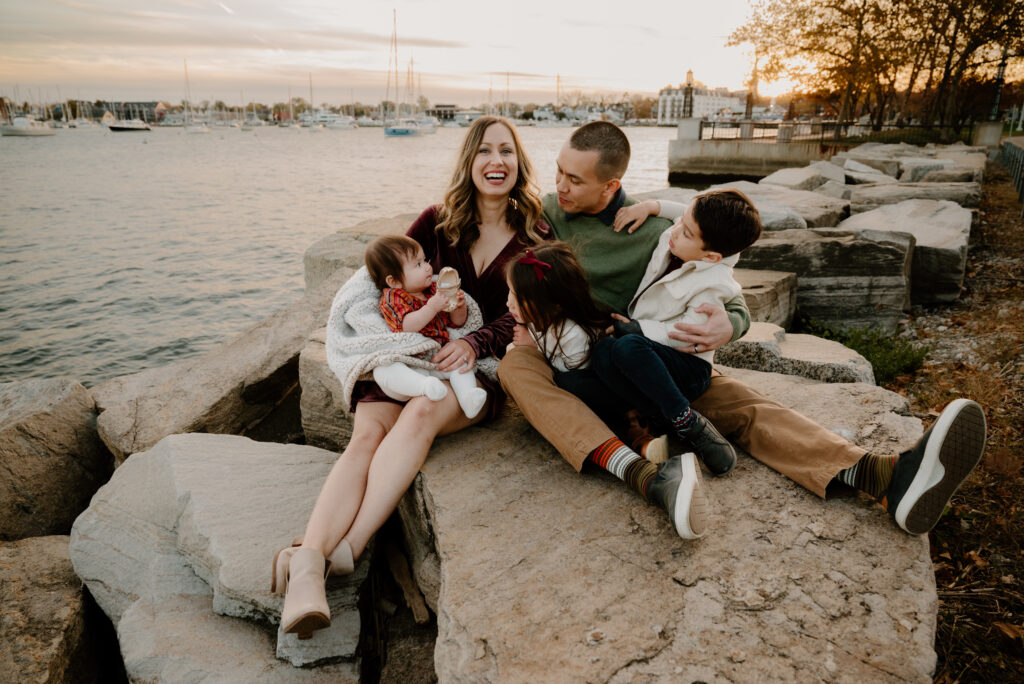 Family sitting together by the water in Annapolis, snuggling close while mom laughs during a relaxed lifestyle photo session.